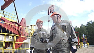 Two man oil workers in orange helmets walking and talking near oil pump jacks and equipment for wellhead connection oil