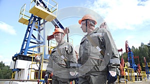 Two man oil workers in orange helmets walking and talking near oil pump jacks and equipment for wellhead connection oil