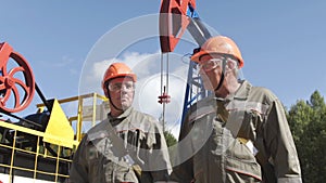 Two man oil workers in orange helmets walking and talking near oil pump jacks. Oil engineers overseeing site of crude