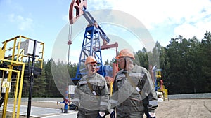 Two man oil workers in orange helmets walking and talking near oil pump jacks. Oil engineers overseeing site of crude