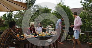 Two Man Taking Food From Barbecue People Sitting At Table Young Friends Group Gathering On Summer Terrace Having Party