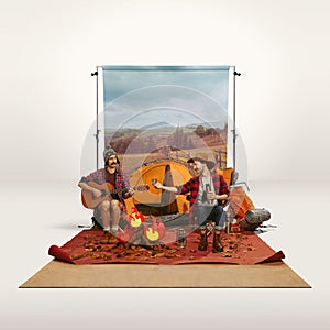 Two mates, men recreating camping activity over grey background with nature wallpaper. Playing guitar, drinking hot tea