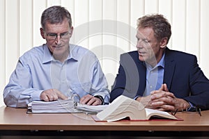Two mature businessmen or partners discussing a project sitting at a desk with folder and book