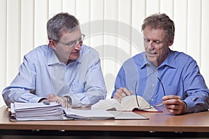 Two mature businessmen or partners readin in a book sitting at a desk with folder and book