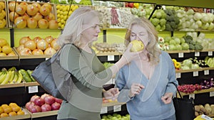 Two mature Caucasian housewives smelling fresh yellow pear in grocery and talking. Confident women discussing fresh