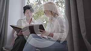 Two mature Caucasian women looking photos sitting on the windowsill. Senior female friends sharing memories at home