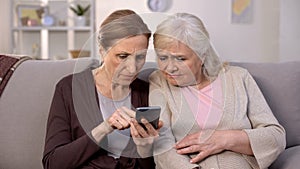 Two mature ladies learning use smartphone sitting home sofa, old age technology