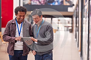 Two Mature Male Students Or Teachers With Digital Tablet Walk Through Hall Of College Building