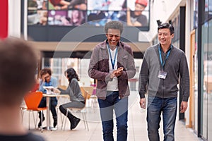 Two Mature Male Students Or Teachers Walking Through Communal Hall Of Busy College Campus Building