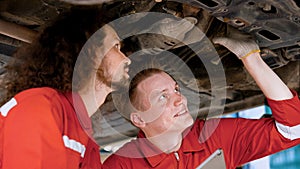 Two mechanic Caucasian man in large garage, Inspecting parts under raised car come in repair problematic part, male mechanic