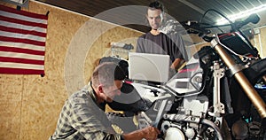 Two mechanic guys are disassembling and inspecting a motorcycle. A guy in a gray T-shirt looks at a laptop screen in a