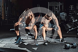 Two men bodybuilders execute exercise with dumbbells for biceps sitting in dark gym