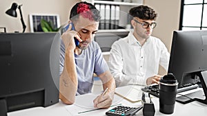 Two men business workers using computer talking on smartphone at office