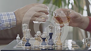 Two men clinking their glasses with whiskeyover a chess board. The beautiful chess set with silver inserts standing on