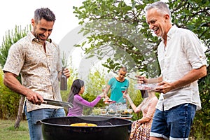 Two men cooking food on grill. Outdoor garden barbecue party. Three women enjoying wine on background.