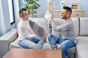 Two men couple fighting with cushion sitting on sofa at home