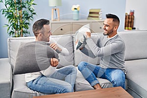 Two men couple fighting with cushion sitting on sofa at home