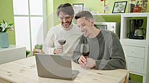 Two men couple having video call drinking glass of wine at dinning room