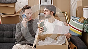 Two men couple talking on smartphone unpacking cardboard box at new home