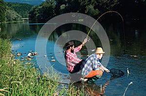 Two men friends fishing. Flyfishing angler makes cast, standing in river water. Old and young fisherman.