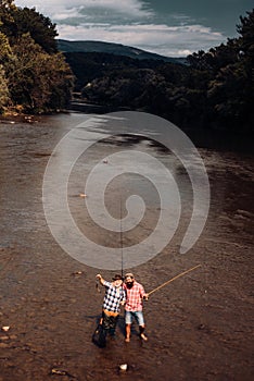 Two men friends fishing. Flyfishing angler makes cast, standing in river water. Old and young fisherman.