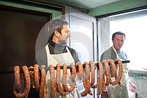 Two men hanging homemade raw sausages on wooden stick.