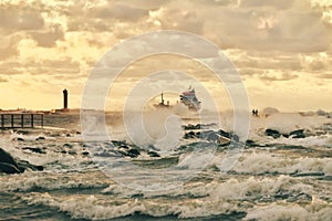 Two men with kite boards standing on the pier and watching as a vessel is arriving to a harbor, breaking through the stormy waves
