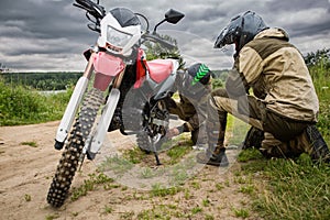 Two men in motorcycle gear checking rear wheel of dirtbike.