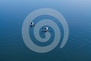 Two Men Paddling Away From Camera On Blue Water With Wake