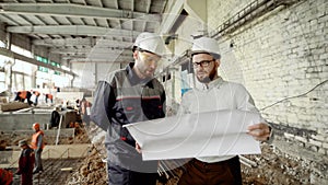 Two men in protective helmets are working together in construction area with workers welding in the background. Foreman