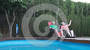 Two men relaxing sitting outdoors in a garden near a swimming pool and a dog.