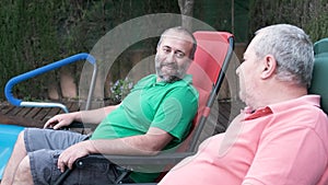 Two men relaxing sitting outdoors in a garden near a swimming pool and a dog.