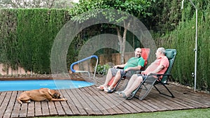 Two men relaxing sitting outdoors in a garden near a swimming pool and a dog.