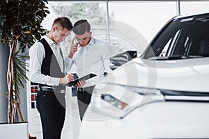 Two men stand in the showroom against cars. Close-up of a sales manager in a suit that sells a car to a customer. The