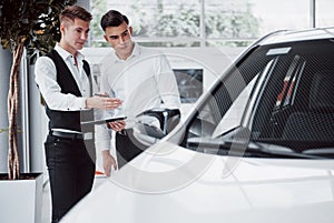 Two men stand in the showroom against cars. Close-up of a sales manager in a suit that sells a car to a customer. The