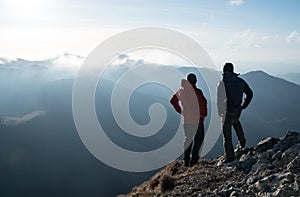 Two men standing standing with trekking poles on cliff edge and looking at sunset rays over the clouds. Successful summit concept