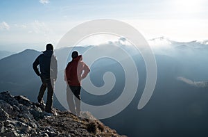 Dva muži stojící s trekkingovými holemi na okraji útesu a při pohledu na paprsky slunce nad mraky. Úspěšný koncept summitu