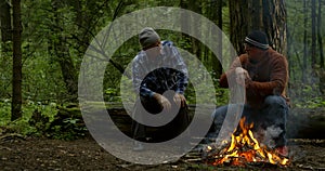 Two men are talking sitting on log in forest near flaming campfire in dusk time