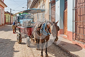 Two men work on flat tire on blue truck behind horse and wagon in Cuban town street