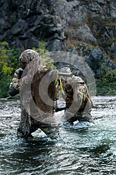 Two mercenary military soldiers during a sabotage mission behind enemy lines - he stands knee-deep in the river and aims