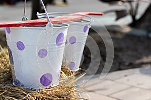 Two metal tin buckets painted with white paint with purple circles and a red rocker on them stand on a haystack