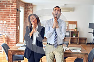 Two middle age business workers standing working together in a meeting at the office begging and praying with hands together with