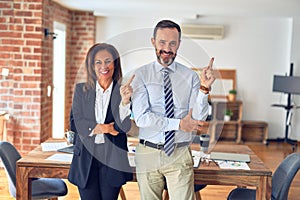 Two middle age business workers standing working together in a meeting at the office with a big smile on face, pointing with hand