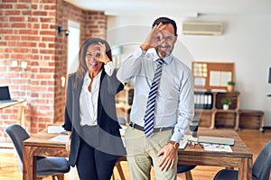 Two middle age business workers standing working together in a meeting at the office doing ok gesture with hand smiling, eye