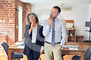 Two middle age business workers standing working together in a meeting at the office with hand on chin thinking about question,