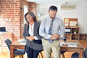 Two middle age business workers standing working together in a meeting at the office with hand on stomach because indigestion,