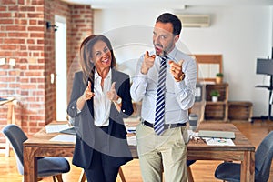 Two middle age business workers standing working together in a meeting at the office pointing fingers to camera with happy and