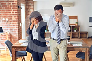 Two middle age business workers standing working together in a meeting at the office with sad expression covering face with hands