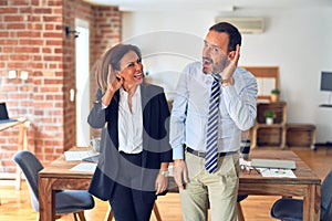 Two middle age business workers standing working together in a meeting at the office smiling with hand over ear listening an