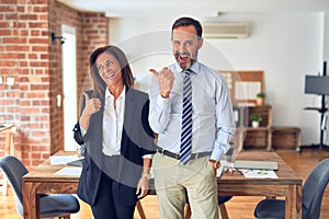 Two middle age business workers standing working together in a meeting at the office smiling with happy face looking and pointing